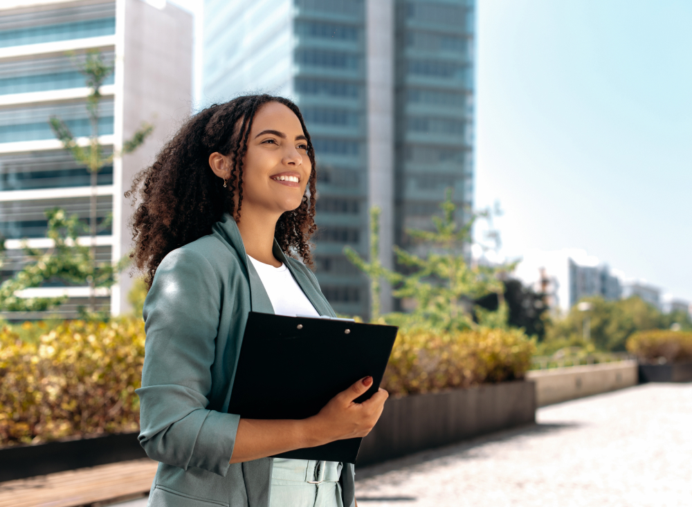 Uma jovem negra de cabelos encaracolados pretos está em frente a um prédio corporativo. Ela segura uma pasta de anotações de executivo. Ela veste camisa branca, calça cinza, casaco formal e olha para frente, passando a ideia de muita determinação, foco no futuro e em busca de suas conquistas.