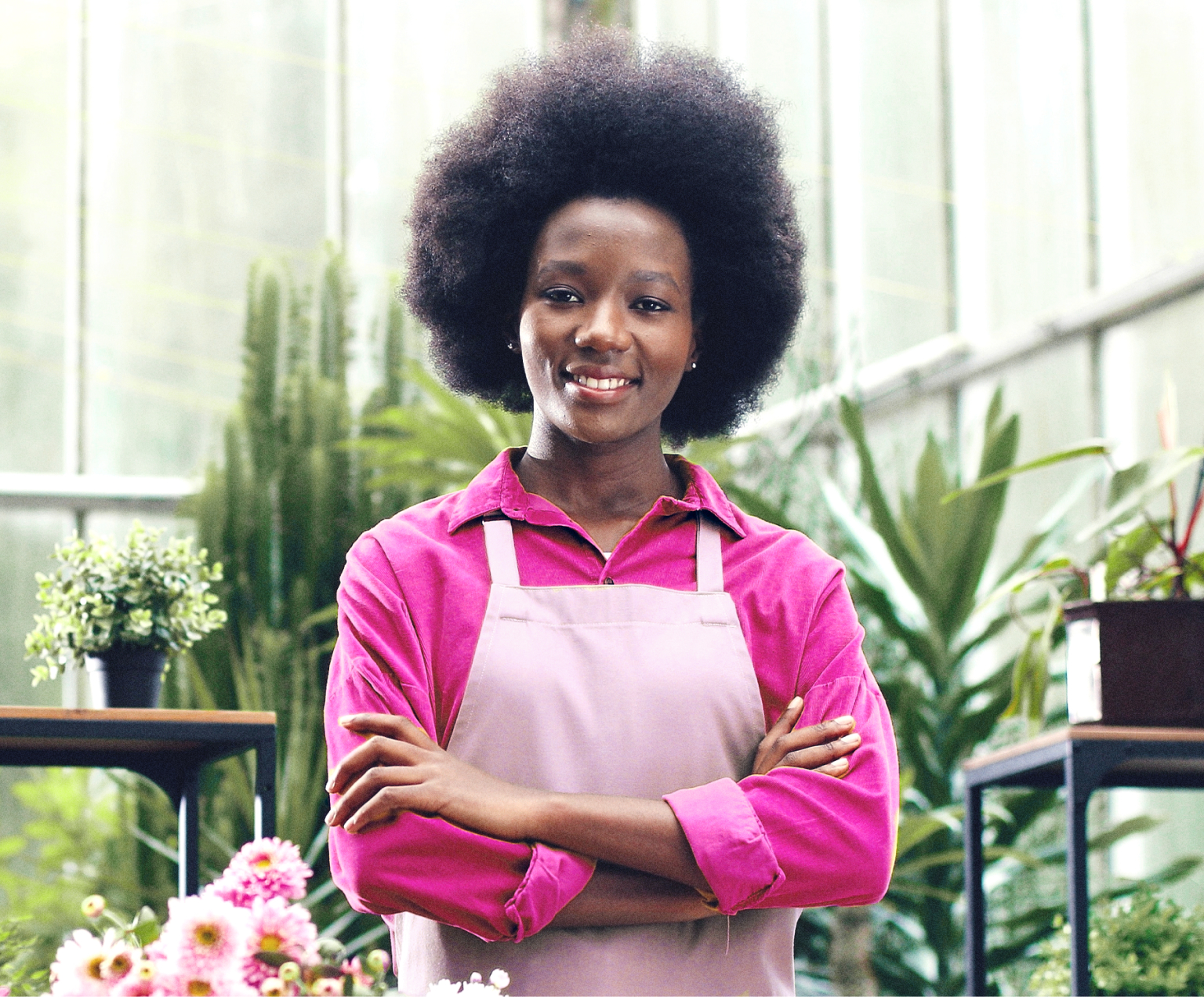 Imagem de uma mulher preta, empresária e empoderada em sua floricultura. Ela está com braços cruzados, sorridente e passando muita confiança e tranquilidade.